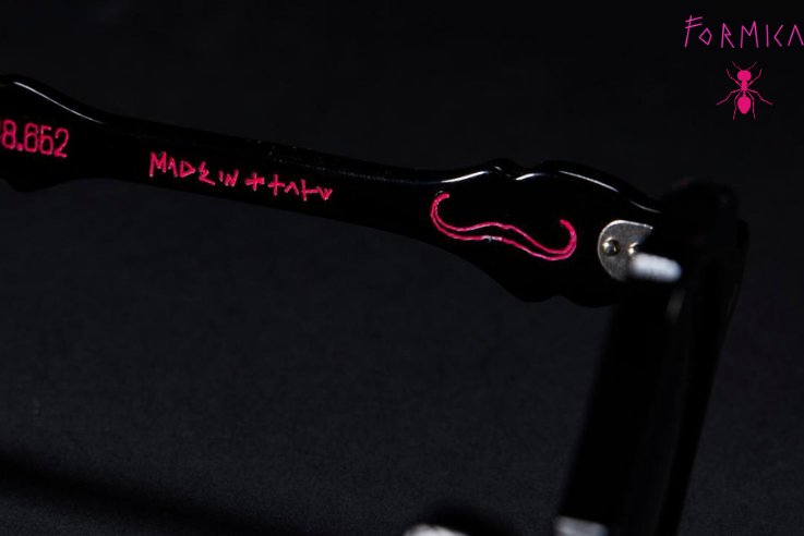 Close-up of black eyeglass arms with red script branding and a red mustache graphic, shown against a dark background with 'MARCEL' and DALI logos visible.