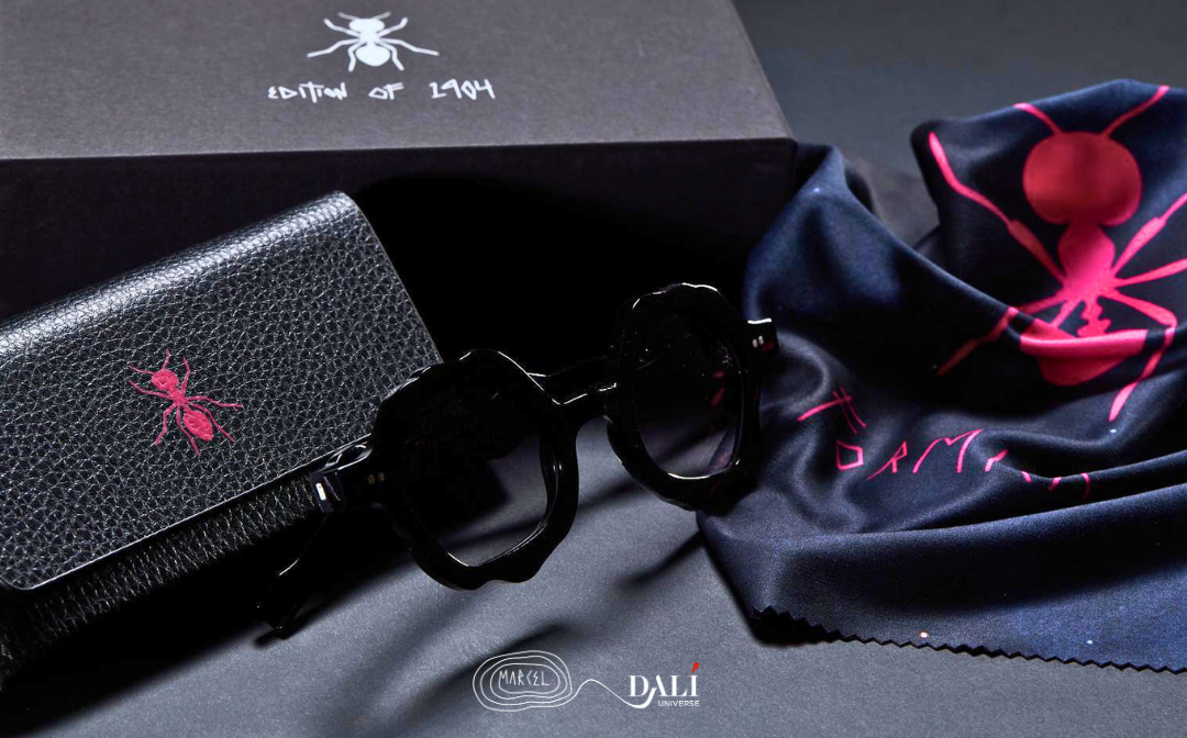 Black round-frame sunglasses displayed with a black textured case, a navy blue silk lens cloth, and a black presentation box with a red ant logo on a dark gray background.