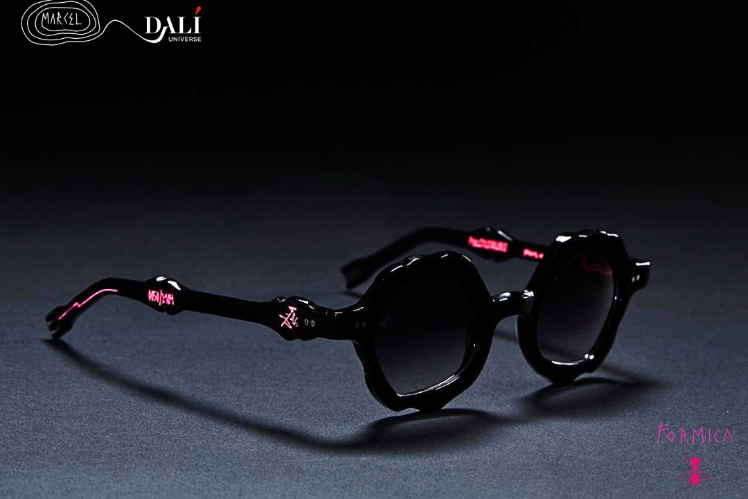 Oversized round sunglasses with thick black frames and dark lenses, featuring pink and white decorative details on the temples, shown on a dark gray surface against a black background. The brand logo 'MARCEL DALI' is visible in the top left corner.