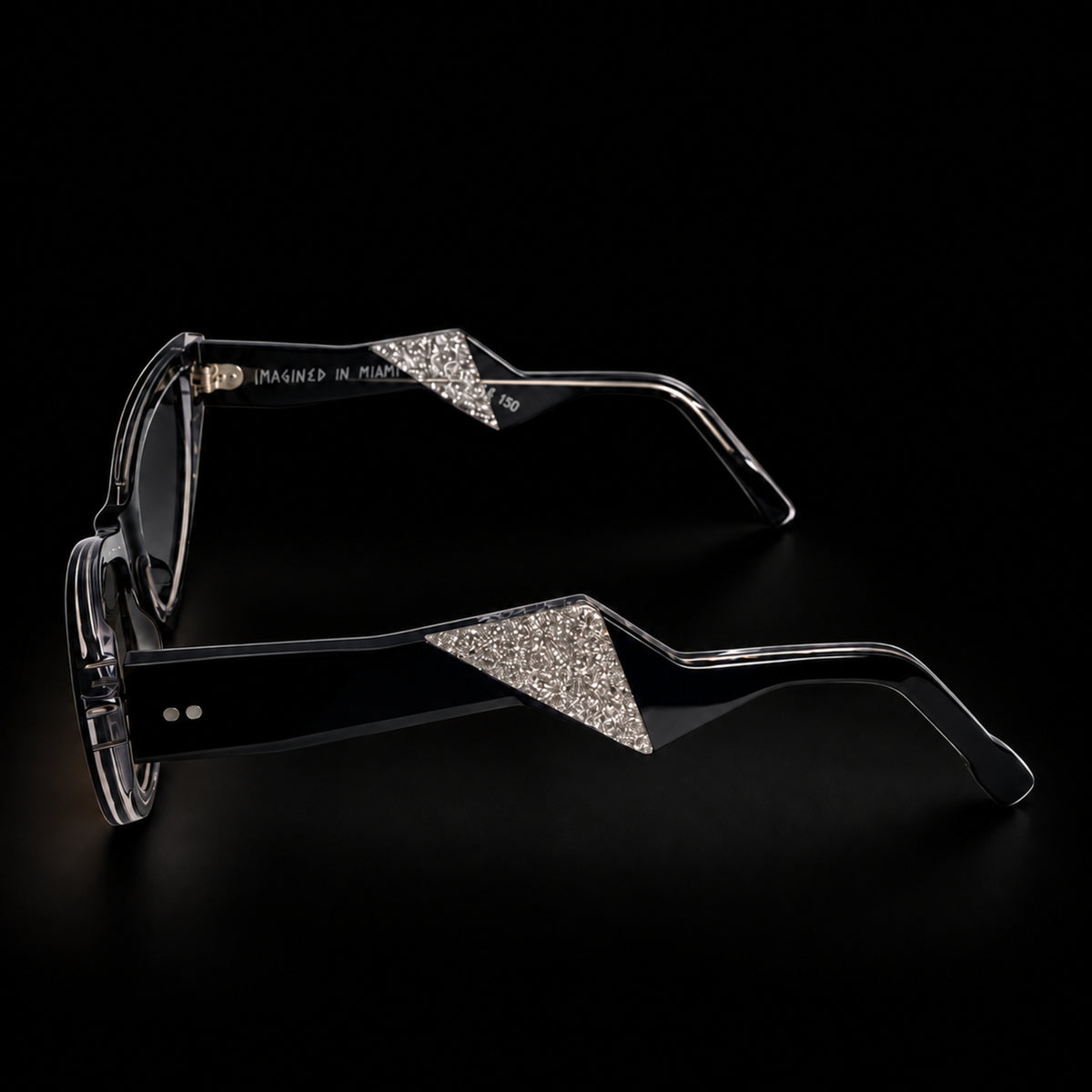 Black cat-eye sunglasses with black gradient lenses and clear temple tips, shown on a black background. The brand name 'MARCEL' is printed on the left lens. Side view.