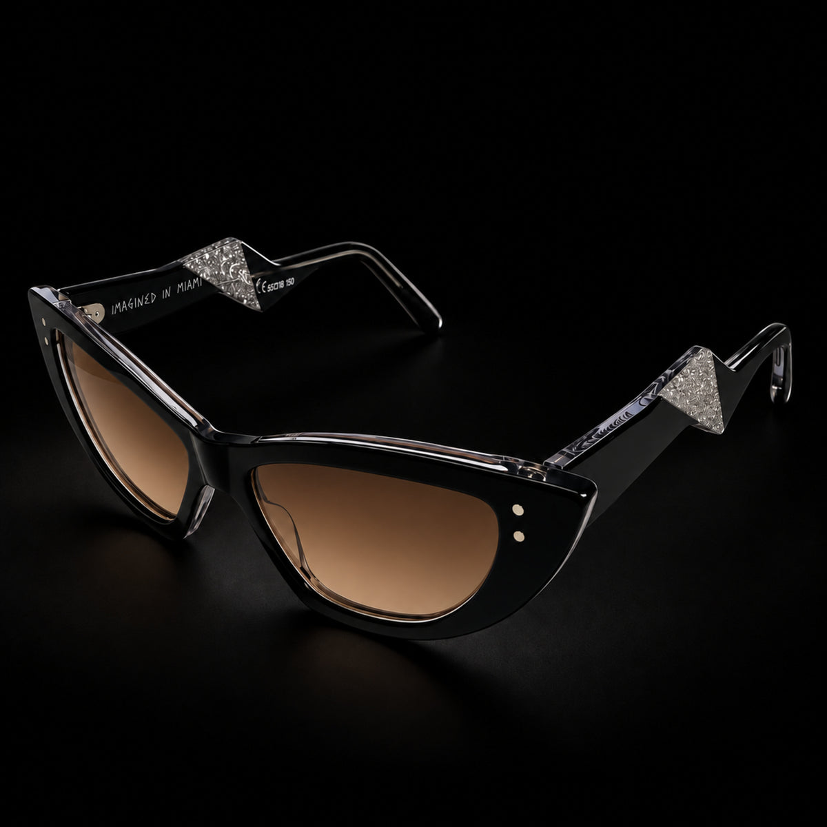 Black cat-eye sunglasses with brown gradient lenses and clear temple tips, shown on a black background. 3/4 view.
