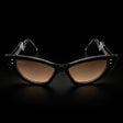 Black cat-eye sunglasses with brown gradient lenses and clear temple tips, shown on a black background. The brand name 'MARCEL' is printed on the left lens. Front view.