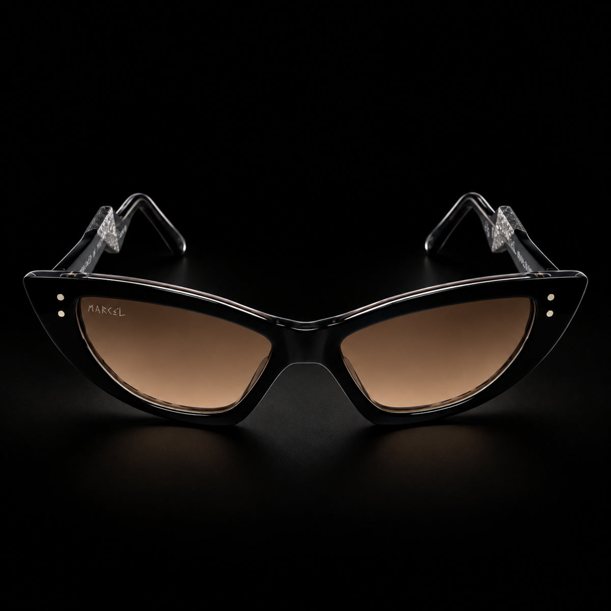 Black cat-eye sunglasses with brown gradient lenses and clear temple tips, shown on a black background. The brand name 'MARCEL' is printed on the left lens. Front view.