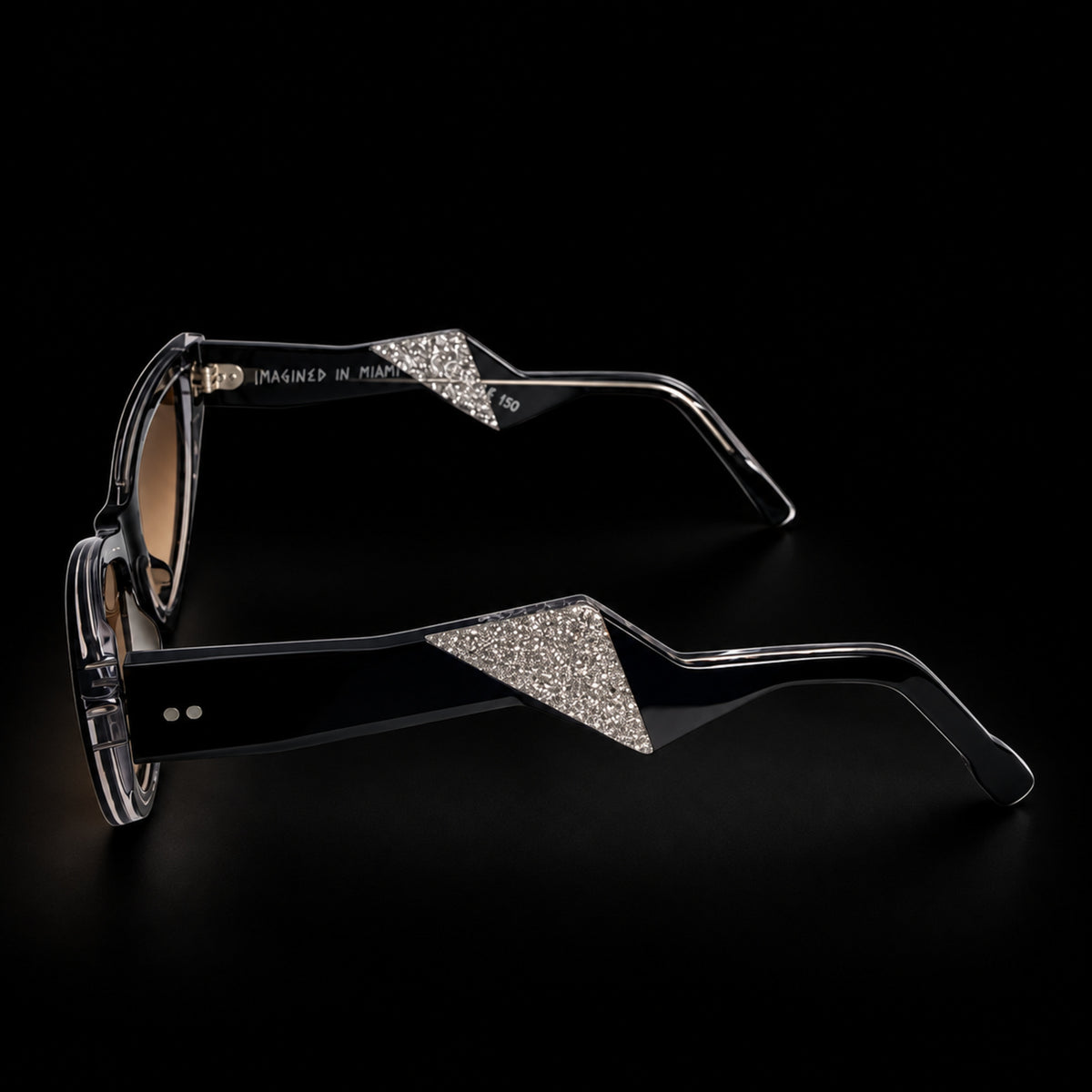 Black cat-eye sunglasses with brown gradient lenses and clear temple tips, shown on a black background. Side view how's silver detailing.