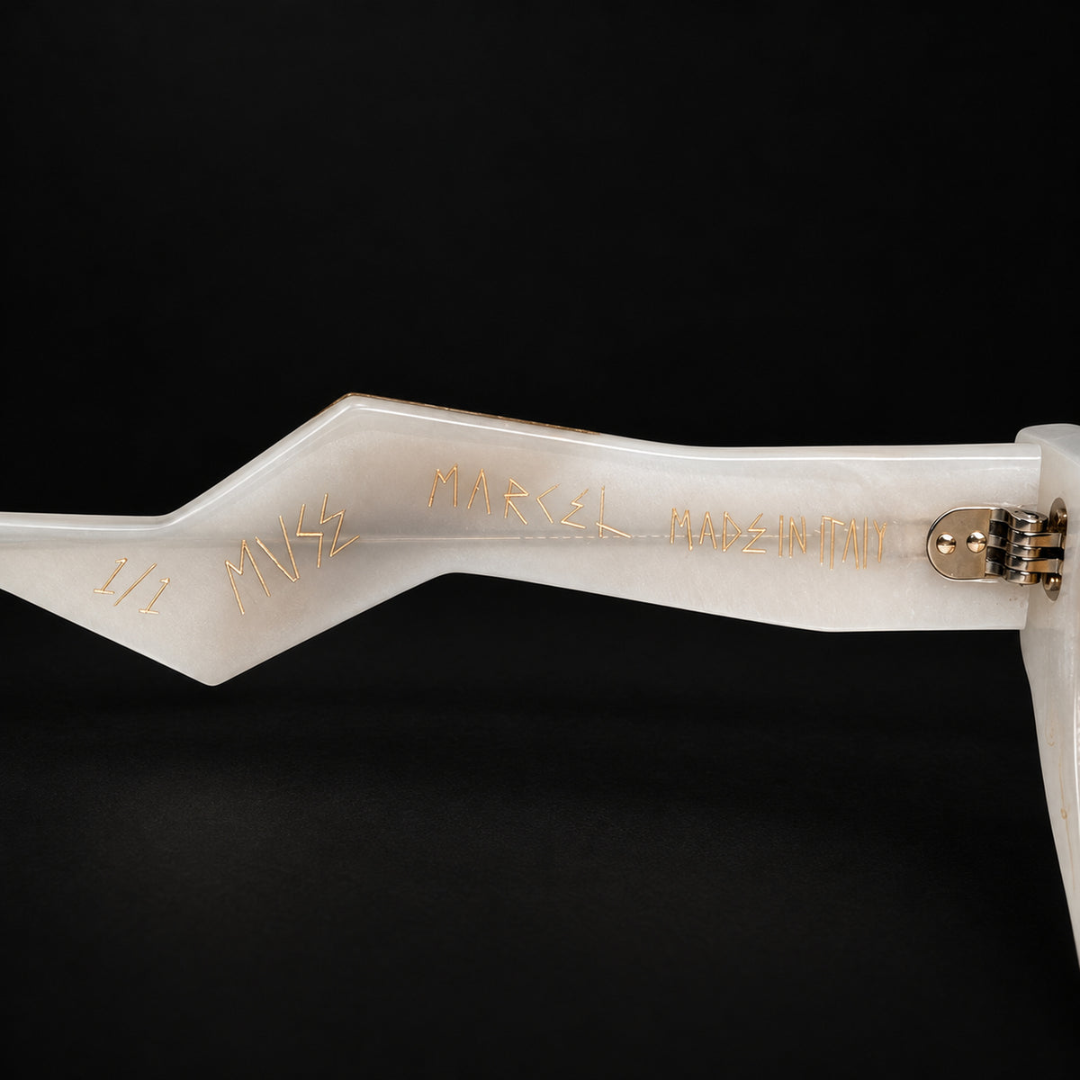 Close-up of a white acetate sunglasses arm with gold foil stamping reading 'MARCEL MADE IN ITALY'  and 'MUSE', set against a solid black background.